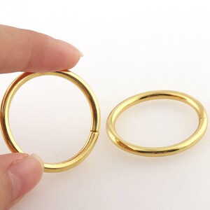 May include: Two gold-colored metal rings. One ring is held by a finger, and the other ring is lying on a white surface. The rings are circular and have a smooth, shiny finish. The rings appear to be new.