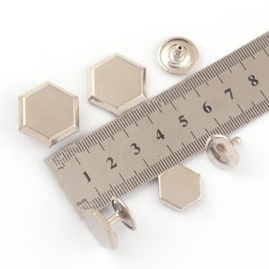 Può includere: Bottoni e chiusure esagonali in metallo argentato. Un righello è posto diagonalmente sull'immagine, mostrando le misure in centimetri. I bottoni hanno una superficie lucida e sono probabilmente utilizzati per abbigliamento o accessori.