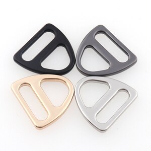 May include: Four metal D-ring buckles in different finishes: black, gunmetal, gold, and silver. Each buckle has a triangular shape with a rounded top and a straight bottom.