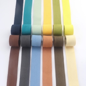 Può includere: Una collezione di nastri in twill a spina di pesce colorati in varie tonalità, tra cui blu navy, turchese, verde chiaro, giallo e oliva. Ogni nastro è arrotolato, con la parte srotolata che si estende verso il basso. Adatti per cucito e artigianato.