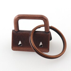 May include: A copper-toned metal key ring with a swivel clasp. The ring is attached to a small, rectangular metal piece with two holes.