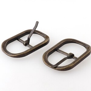 May include: Two antique gold-toned metal belt buckles. Each buckle has a rectangular shape with rounded corners and a single prong.