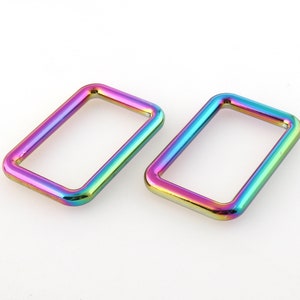 Puede incluir: Dos anillos metálicos rectangulares iridiscentes con esquinas redondeadas. Los anillos muestran un vibrante espectro de colores, incluyendo morado, azul, verde y amarillo. Estos componentes de hardware se utilizan probablemente en la fabricación de bolsos u otros proyectos de artesanía.