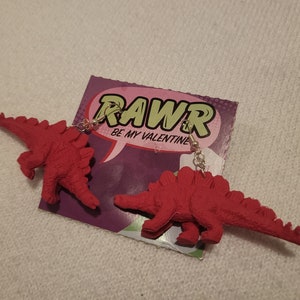 May include: A pair of red dinosaur earrings with silver hooks. The earrings are packaged on a card with the text "RAWR BE MY VALENTINE".