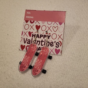 May include: A pair of pink and white skateboard earrings with a heart and X pattern. The earrings are on a Valentine's Day card with the text "Happy Valentine's Day".