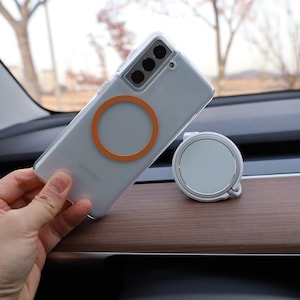 May include: A clear phone case with an orange magnetic ring is attached to a silver smartphone. A white and silver magnetic car mount sits on a wooden dashboard. The phone is held by a hand.