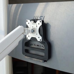 Pode incluir: Um braço de monitor e um suporte de montagem brancos e pretos fixados na parte traseira de um monitor de computador prateado. O suporte tem uma placa de montagem branca e uma estrutura de suporte preta. O monitor tem o logotipo MSI.