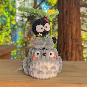 May include: A handmade plush figure of Totoro, a gray creature with a white belly and pink cheeks, topped with a black soot sprite holding a flower. The figures are made of soft, fuzzy material and sit on a wooden surface.