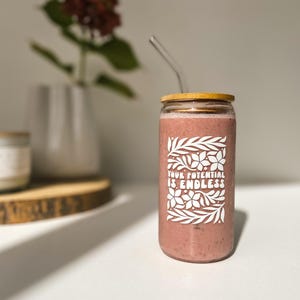 Könnte beinhalten: Ein Glasgefäß mit Bambusdeckel und Strohhalm, gefüllt mit einem rosa Smoothie. Das Gefäß hat ein weißes Design mit dem Text "YOUR POTENTIAL IS ENDLESS" und floralen Mustern.