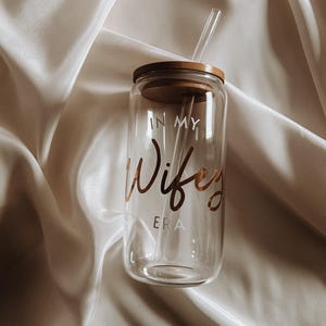 Könnte beinhalten: Ein durchsichtiger Glasbecher mit einem Holzdeckel und einem Strohhalm. Der Becher trägt den Schriftzug "In My Wife Era" in goldener Schrift.