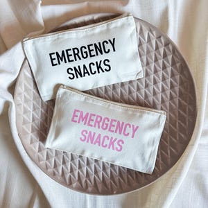 Può includere: Due pochette in tela bianca con la scritta "Emergency Snacks" stampata su di esse. La pochette superiore ha il testo nero e la pochette inferiore ha il testo rosa.