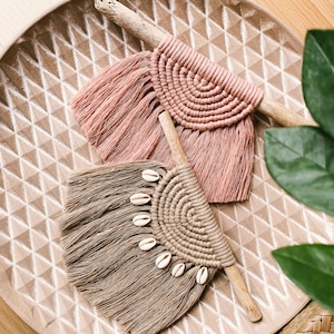 Könnte beinhalten: Zwei Makramee-Wandbehänge mit Fransen, einer in Rosa und einer in Braun mit weißen Muscheln. Die Behänge befinden sich auf einem hellbraunen Tablett mit einem Rautenmuster.