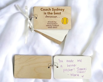 May include: A wooden book with a white ribbon and a softball graphic. The cover reads "Coach Sydney is the best because... A book by: Firecrackers Infinity Softball 2022-2023". A separate card reads "You made me a better pitcher! Thanks Coach! - Maria ♡".