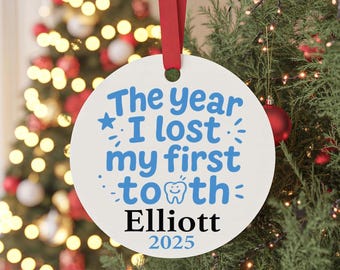 May include: A round white ceramic ornament hanging from a red ribbon. The text reads The year I lost my first tooth in blue, with a small tooth graphic, followed by the name Elliott in black and the year 2025 in blue at the bottom.
