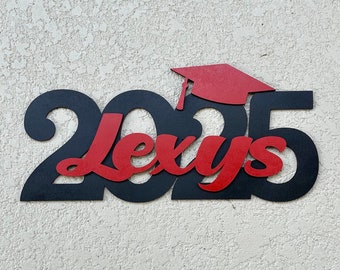 May include: A black and red graduation sign with the text "2025 Lexys" and a red graduation cap. The sign is made of a solid material and is mounted on a textured white wall. The design is a celebratory theme.