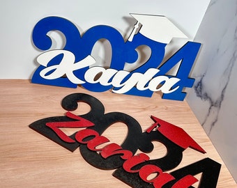 May include: Two graduation signs, one blue with the name "Kayla" and the year "2024" and one black with the name "Zariah" and the year "2024".