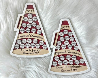 May include: Two red and white megaphone-shaped wooden signs with the text "Regional Champions" and the year "2022". Each sign has a list of names in white lettering on a burgundy background. The sign on the left says "Coach Jackie Somers D12" and the sign on the right says "Coach Stephanie Somers D12".