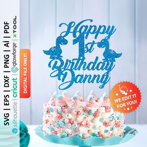 Peut inclure: Décoration de gâteau bleu pailleté avec le texte "Happy 1st Birthday Danny" et des silhouettes de dinosaures. Le gâteau est décoré de glaçage rose, bleu et blanc. Le gâteau est sur un présentoir bleu.