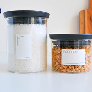 Puede incluir: Dos tarros de vidrio con tapas negras y etiquetas blancas. El tarro de la izquierda contiene sal gruesa y el tarro de la derecha contiene palomitas de maíz. Ambas etiquetas tienen la palabra "PANTRY" impresa.