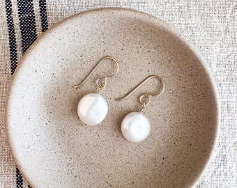 May include: A pair of white pearl earrings with gold hooks, displayed on a beige ceramic dish.