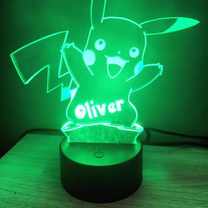 May include: A green LED night light depicting Pikachu, with the name "Oliver" in white. The light sits on a black base with a power symbol. The Pikachu figure is in a cheerful pose, with its arms outstretched.