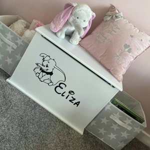 May include: White toy chest with a grey star pattern on the front. The chest is decorated with a black and white sticker of Dumbo the elephant and the name "Eliza".