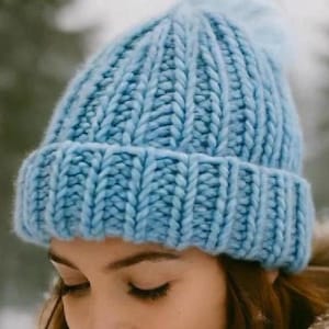 Puede incluir: Un gorro de punto azul claro con pompón, usado con un abrigo de invierno beige con capucha forrada de piel. El gorro está hecho de hilo grueso y grueso, perfecto para el clima frío. El fondo es una escena nevada al aire libre.