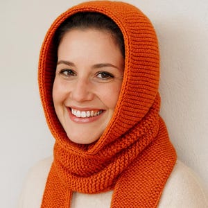 May include: A knitted hood and scarf set in a vibrant orange. The hood frames the face and flows into a scarf. The knitwear is a solid orange colour. The person is wearing a cream-coloured jumper.