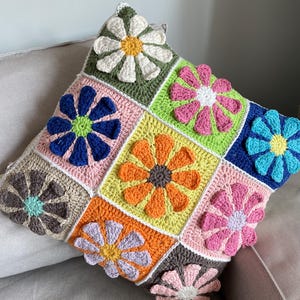 Könnte beinhalten: Ein quadratisches Häkelkissen mit einem Patchwork-Muster aus bunten Blumen. Das Kissen ist aus verschiedenen Farben Garn gefertigt, darunter Weiß, Grün, Rosa, Blau, Orange und Braun. Das Kissen liegt auf einem hellbraunen Sofa.