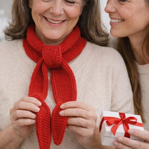 May include: A red knitted scarf is held by a person. The scarf is tied in a knot at the neck and has two long, pointed ends. A small white gift box with a red ribbon is held nearby. The scarf is a cozy accessory.