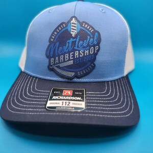 May include: A blue and white trucker hat featuring the text "Next Level Barbershop EST. 2023" and a barber shop logo. The hat has a dark blue brim and a Richardson tag.