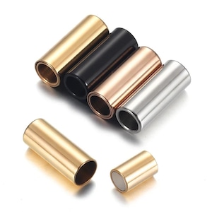 Può includere: Una collezione di slide per chitarra in metallo color oro, nero, oro rosa e argento. Le slide cilindriche sono mostrate su una superficie bianca. Una slide dorata è mostrata separatamente, insieme a una slide dorata più piccola con un magnete.