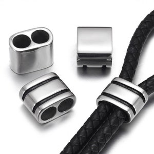 Puede incluir: Tres tapones de extremo de cordón de metal plateado, dos con agujeros dobles y uno con un agujero simple, con líneas negras alrededor de los bordes. Los tapones de extremo de cordón se muestran en un cordón de cuero negro.