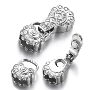 May include: A set of three silver-colored magnetic clasps. The clasps have a floral design with a central circle and petal-like shapes around the edge. One clasp is open, revealing a magnet.
