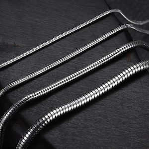 May include: Four silver snake chains of varying lengths and widths, displayed on a black surface.