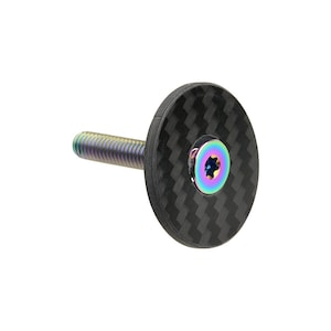 Puede incluir: Una tapa de potencia de bicicleta de fibra de carbono negra con un tornillo de color arcoíris. La tapa tiene una superficie texturizada.