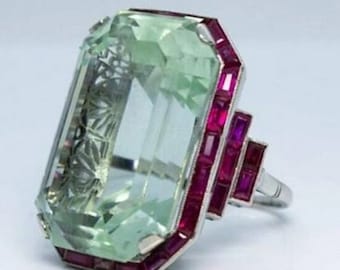 May include: A silver ring with a large, light green emerald-cut gemstone surrounded by a border of small, rectangular ruby stones.