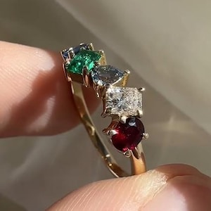 Könnte beinhalten: Ein goldener Ring mit einer Reihe farbiger Edelsteine. Der Ring zeigt eine Abfolge von blauen, grünen, grauen, weißen und roten Edelsteinen. Die Edelsteine sind in einem goldenen Band gefasst.