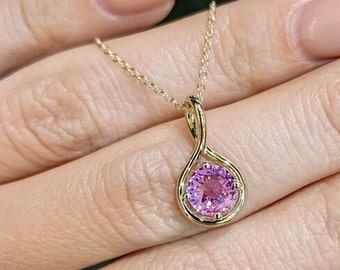 May include: A gold necklace with a pendant holding a round, pink gemstone. The pendant has a delicate, swirling design. The fine chain enhances the pendant's elegance. A piece of jewellery.
