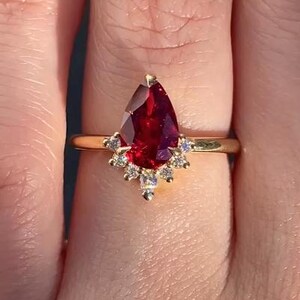 May include: A gold ring featuring a pear-shaped, deep red gemstone as the centerpiece. The gemstone is surrounded by smaller, clear accent stones. The ring's band is a thin, polished gold.