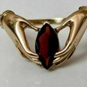 May include: A gold ring with two hands holding a large, red, marquise-shaped gemstone.