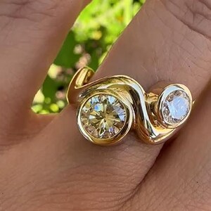 May include: A gold ring featuring two round-cut diamonds. The larger diamond has a champagne hue, while the smaller one is clear. The ring's band has a unique, flowing design.