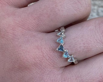 May include: A silver ring featuring a series of triangular, light blue gemstones. The ring is set with multiple, evenly spaced, triangle-shaped stones. The ring is worn on a finger, showcasing the design and color of the gemstones.