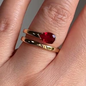 Peut inclure: Une bague dorée ornée d'une pierre précieuse rouge foncé en forme de goutte d'eau. Le corps de la bague s'enroule en spirale autour du doigt. La pierre est sertie à griffes. La bague est portée sur un doigt.