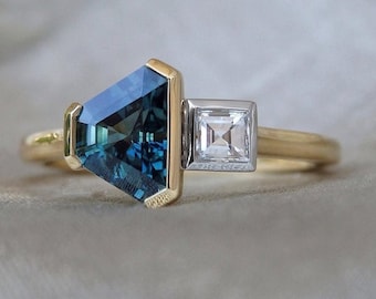 May include: A gold ring with a blue gemstone and a smaller, clear diamond. The ring is set in a modern, geometric style.
