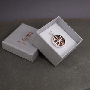 Op de afbeelding: Een zilveren kompas hanger met een bruine houten inlay in een grijze geschenkdoos. De doos heeft de tekst "AMORE & AVVENTURA" erop gedrukt.