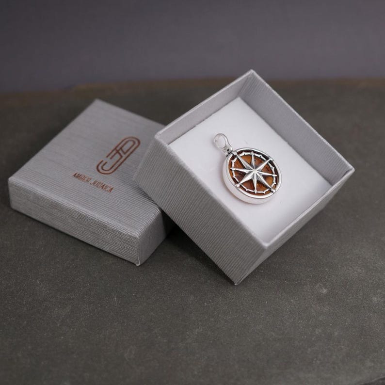 Op de afbeelding: Een zilveren kompas hanger met een bruine houten inlay in een grijze geschenkdoos. De doos heeft de tekst "AMORE & AVVENTURA" erop gedrukt.