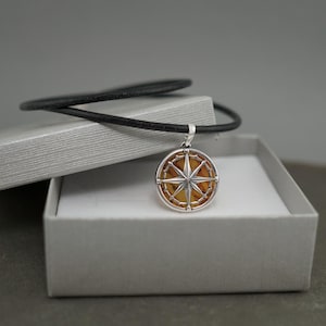 Op de afbeelding: Een zilveren kompas hanger met een bruin amberen hart, op een zwart leren koord, in een grijze geschenkdoos.