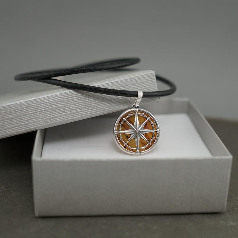Op de afbeelding: Een zilveren kompas hanger met een bruin amberen hart, op een zwart leren koord, in een grijze geschenkdoos.