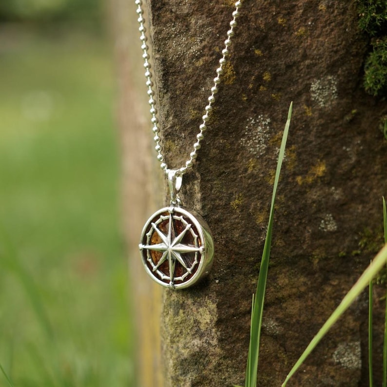 Op de afbeelding: Een zilveren ketting met een ronde kompas hanger. Het kompas heeft een zilveren frame en een bruin centrum met een zilveren ster.